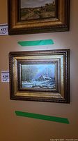 Photo showing the framed oil painting on the wall under a larger painting, with green tape below and the lot tag visible.
