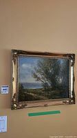 Framed oil painting hung on wall showing the entire painting of a landscape with trees, water and sky. Ornate gold frame is visible.