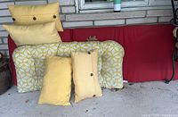 Three white and yellow floral patterned rectangular cushions with two yellow pillows with button closures, and one plain yellow pillow, arranged on red outdoor seating against wall.