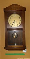 Front view of wooden pendulum wall clock showing clock face with Arabic numerals and visible pendulum inside wooden case.