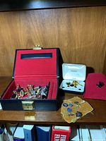 Wide view showing black box with red velvet lining and multiple pins and cufflinks inside, white branded case with golden cufflinks, red pouch with a pair of black cufflinks, and fabric cloth with various pins.