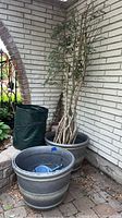 Two large gray plastic planters placed near a white brick wall and stone floor, one containing a faux tree and the other empty. Green collapsible yard waste bag visible behind them.