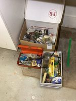 Overview photo showing three containers with assorted plumbing hardware, soldering supplies, and tools including a red metal toolbox and cardboard boxes.