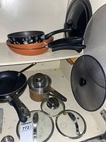 Overall view of the assortment of pots, fry pans, glass lids, and mesh splash screen stacked on shelf.