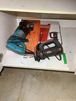 Two electric power tools on a white shelf including a teal reciprocating saw and black/red Jobmate cut-out tool with their boxes behind them.