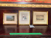 Three vintage framed prints with gold frames displayed together against a dark wood background, showing overall condition and size.