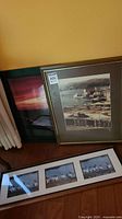 Three framed prints leaning against a wall, showing one nautical harbor scene, one sunset, and one pelican triptych.