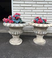 Two cream resin planter urns with scalloped rims and bulbs at the base, each filled with bright pink faux flowers and green leaves, set on a grey pavement by a white brick wall.