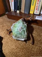 Semi precious green crystalline stone cluster resting on wooden tree branch stand with bookshelf background.
