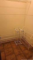 View of three different white metal shoe racks in a corner of a room with tiled floor, showing all racks with some corrosion and wear.