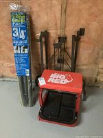 Photo of Torin Big Red rolling shop stool with padded red vinyl seat, metal frame base, and black base tray on caster wheels, positioned next to two metal table legs and the unopened pipe insulation tubes.