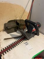 Side view of Craftsman cordless hedge trimmer and metal folding entrenching tool with carrying case on white surface.