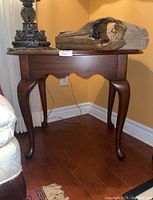 View of the oval wooden vintage side table showing the front and one side. Table has four curved legs and a scalloped apron beneath the top. The wood has a dark finish and the top appears smooth and polished. Some decorative items rest on top, not included in the lot.