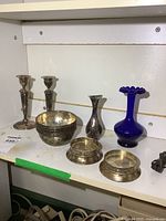 Full view of the blue glass bud vase, silver plated bowls, candle holders and coasters on white shelf with visible tarnish