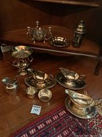 Overall view of silver plated dining pieces arranged on wood shelves including gravy boats, urn, drink shaker, tray, pitcher, creamer, sugar bowl. Tarnish visible on surfaces.