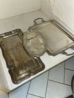 Three tarnished silver plated serving trays on a white surface, shows overall condition and shape variations of trays including oval, rectangular with handles, and smaller tray.
