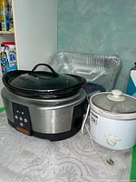Large stainless steel Crock Pot slow cooker with glass lid and removable bowl, alongside smaller white Toshiba slow cooker with dial control and lid knob. Several aluminum foil toasting pans behind.