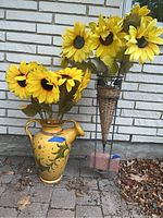 Front view of painted yellow watering pot with floral designs and a bunch of faux sunflowers inside, sitting outdoors on a stone surface.