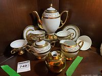 Full view of the white porcelain tea set with gold floral accents and gold trim including teapot, six cups with matching saucers, cream and sugar pots all displayed on wooden surface.