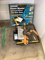 Front view of tile wet saw with visible diamond cutting wheel, orange safety guard, power cord, measurement scales, instruction manual, and original box showing product details.