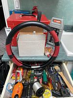 Drawer and countertop showing an assortment of hand tools including screwdrivers, pliers, wrenches, a clamp, socket set parts, a small red metal toolbox, a black and red steering wheel cover, a grout container, and a bottle of leather cleaner.