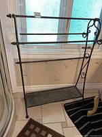 Front view of black and gold metal towel rack positioned in front of window, showing two horizontal bars and lower shelf.