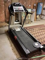 Full treadmill view showing the entire unit folded down on a basement floor with a carpet underneath, control panel, and running belt visible.