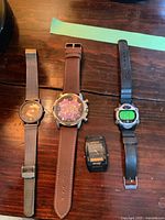 Photo showing four watches side by side on wooden surface: Skagen watch with metal strap, Expedition with plastic strap, Daniel Hechter with synthetic strap, Casio watch without strap