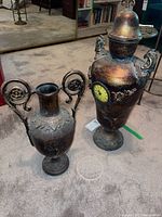 Photo showing both urns positioned on carpet floor, highlighting size difference, texture, and ornate handles.