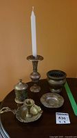 Photo of the four etched brass items on table: candlestick holding white candle, lidded container, rose bowl, ashtray