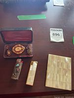 Lot contents on table showing cigarette case, lighters, vintage brooch and earrings case.