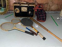 Photo showing entire lot including two wooden tennis rackets, badminton racket, wooden baseball bat, plaid sports bag, and vintage suitcase with travel stickers