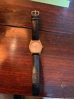 Front view of Rotary gold-tone wristwatch with black leather band laid flat on wooden surface.