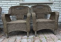 Pair of brown rattan wicker armchairs placed on a brick patio in front of a white brick wall.