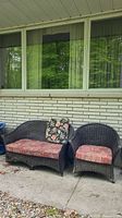 Wicker loveseat and chair in front of window on concrete porch, showing overall set with cushions and accent pillow.