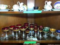 Display of twelve red wine and sherry glasses with gold decorative bands and five smaller multicolored glasses on a glass shelf.
