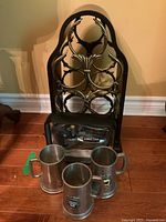 Photo of wooden wine rack holding no bottles, Kirkland corkscrew set in case in front, and three pewter beer steins placed on floor, showing overall lot contents.