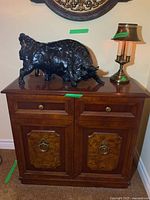 Front view showing wood Craftline cabinet with two drawers, two doors with burl wood panels and brass hardware, black bull sculpture and table lamp on top.