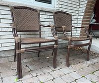 Two brown metal framed chairs with woven seats and backrests, arranged on a tiled patio against a white brick wall.