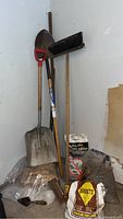 Shovel, broom, metal pipe, and bags of concrete materials in corner of garage.