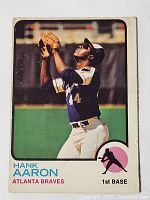 Front photo of 1973 OPC Hank Aaron baseball card showing Hank Aaron in baseball uniform catching a ball with green background and card markings.