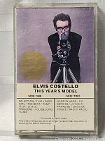 Front cover of vintage Elvis Costello cassette tape This Year's Model inside clear plastic case. Image of singer with camera and track listings visible.