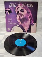 Photo showing vinyl record laid flat with cover propped behind, vinyl has blue center label and shows minimal wear.