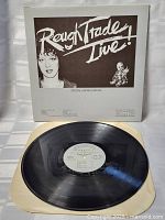 Full view of the vinyl record partially out of its white inner sleeve showing the black vinyl and centered white label with track information.