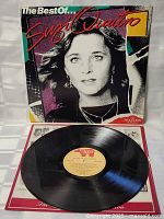 Vinyl record with beige center label placed on red and white paper insert, with album cover behind it showing signs of age wear.