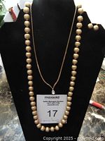 Gold-tone bead necklace displayed on a black bust with a pair of matching gold-tone stud earrings placed on the upper corner of the bust.