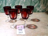 Five vintage cranberry glass goblets displayed from side view, showing their round bowl shape and clear stems on a beige surface.