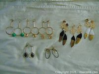 Photo showing 8 pairs of handmade earrings on a light cloth background, including gold hoops with beads and charms, small black stone studs, and feather earrings.