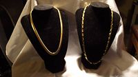 Two gold plated necklaces displayed on black velvet stands: one thick flat herringbone necklace and one chain with elongated oval links.