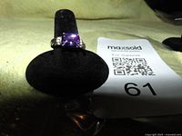 Ring displayed on black velvet pad with tag showing lot number 61. Purple cubic zirconia stone, square cushion cut, mounted on band with side grooves.